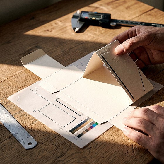 Hands lifting a vellum overlay off a printed structural dieline, steel rule and calipers alongside